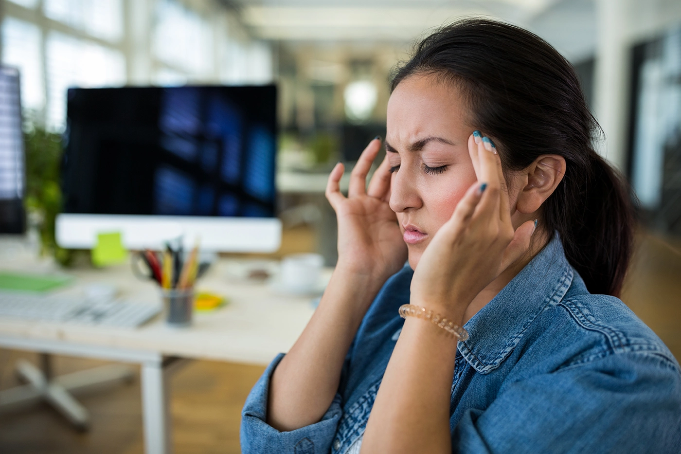 Woman holding head in office suffering from migraine, searching for effective migraine treatment.